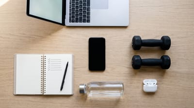 Overhead view of a laptop, a phone, two small black dumbbells, an open planner with a ticked checklist, a water bottle and earbuds on a wooden desk